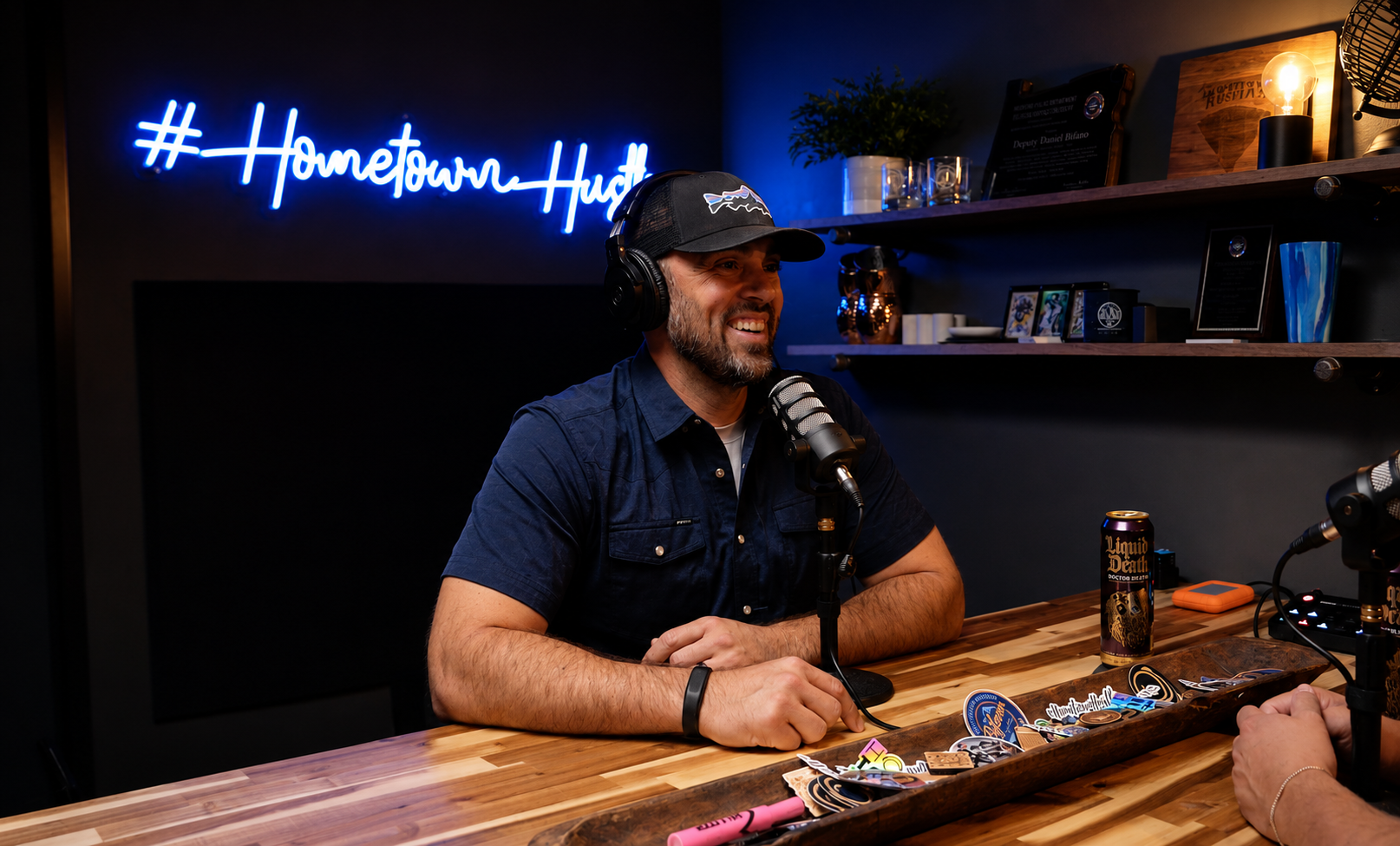 Daniel Bifano at the mic under the blue Hometown Hustle neon sign in the Grants Pass studio